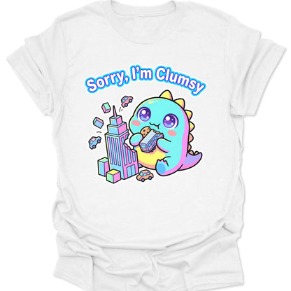 White unisex adult t-shirt featuring a pastel chibi kaiju eating a bus with "Sorry, I'm Clumsy" text in bubbly letters.