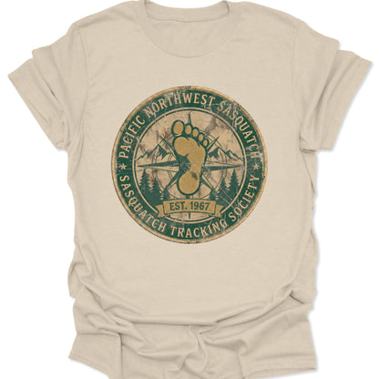 Natural unisex adult t-shirt featuring a retro Pacific Northwest Sasquatch badge with a large yellow footprint and compass rose.